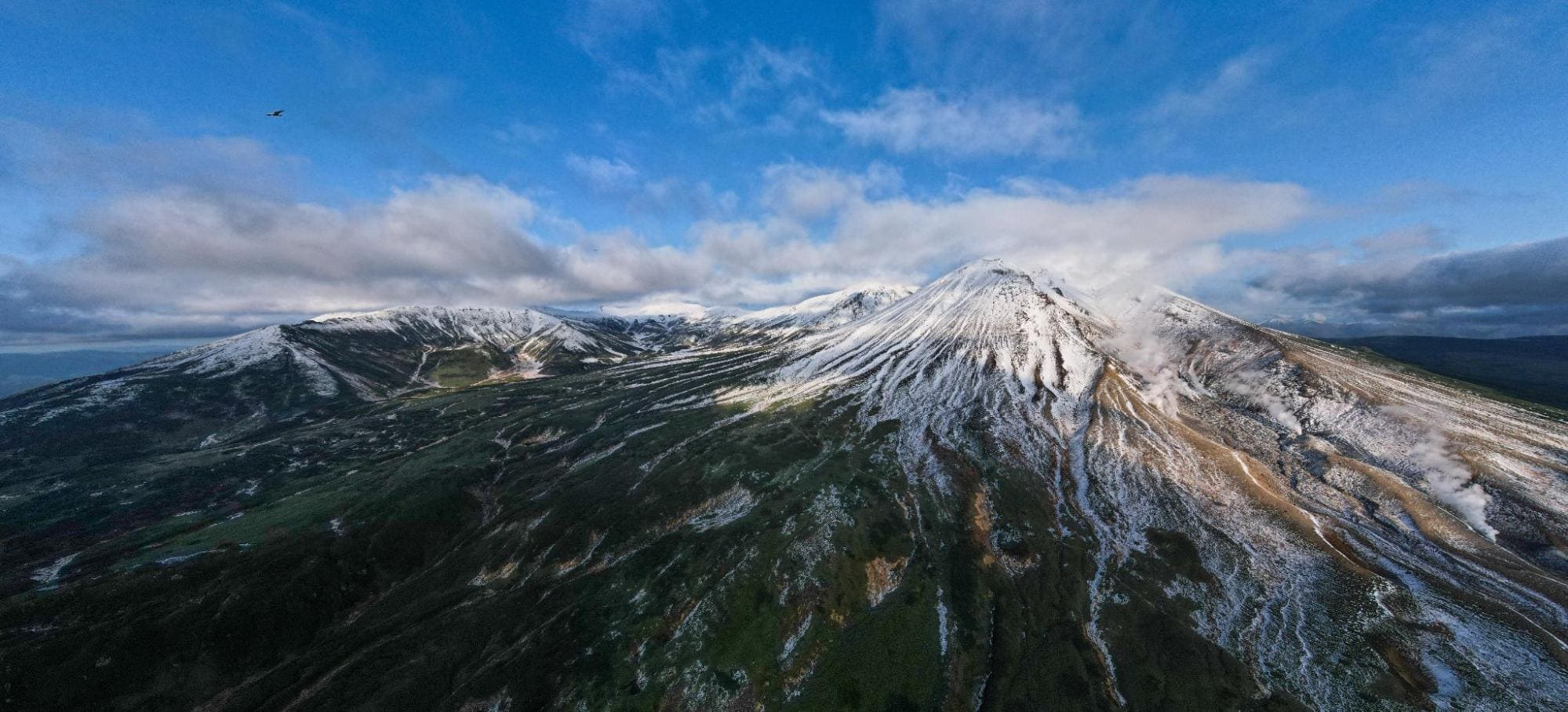 Aerial drone shot of Mount Asahidake with a fresh snowfall, a bird flying overhead, and clouds partially covering the peak.